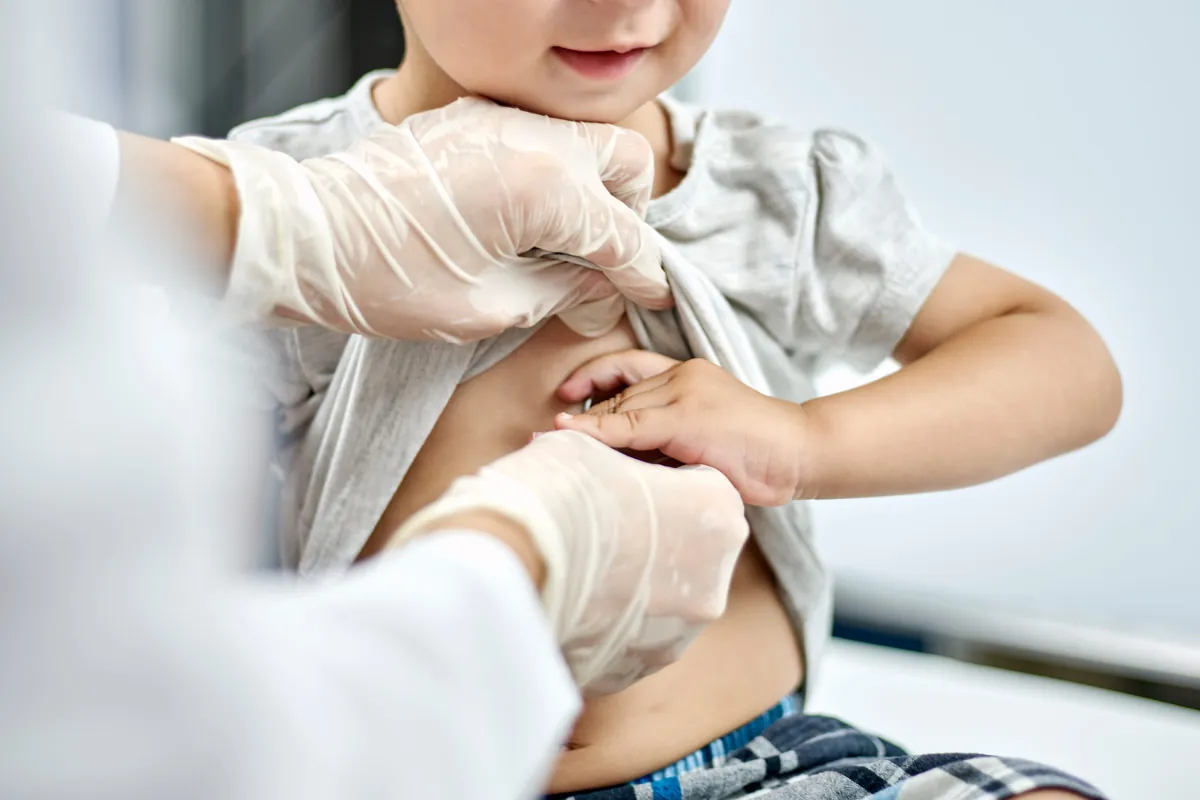 Gloved pediatric provider gently examining a smiling toddler’s tummy