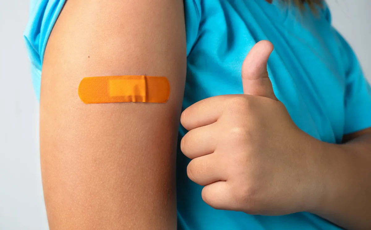 Child in a blue shirt giving a thumbs up next to an orange bandage on his arm