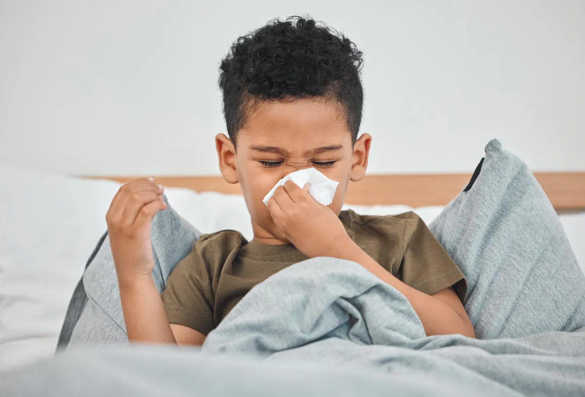 Young boy sitting up in bed blowing his nose while sick with a cold